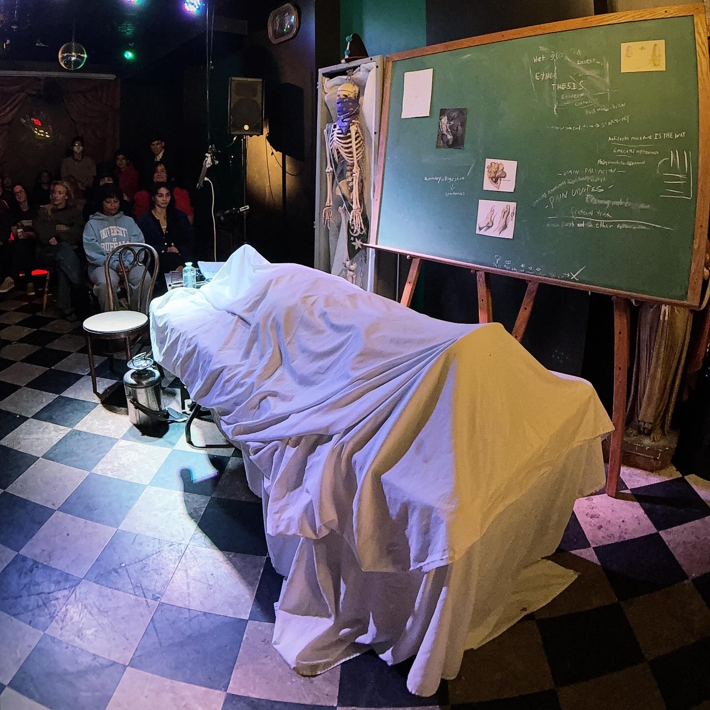 A corpse lies under a sheet on an operating table. Behind the table is a chalkboard with various medical diagrams pasted on, and a tray of medical implements.